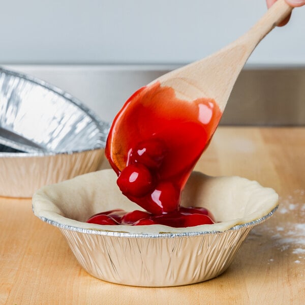 A spoon pouring red cherry pie filling into a D&W Fine Pack round foil pie tin.