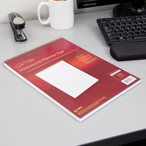 A brown Ampad planner pad on a desk with a white notebook, keyboard, and stapler.