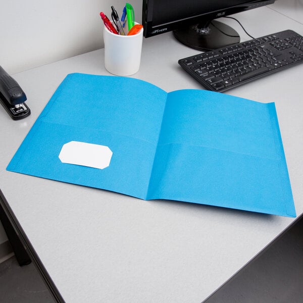 A light blue Oxford 2-pocket paper folder on a desk with a computer and pens.