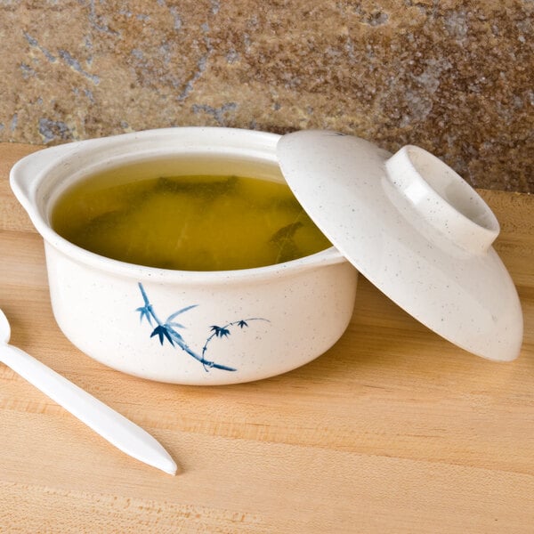 A blue bamboo Thunder Group melamine miso bowl with broth, a lid, and a spoon on a table.