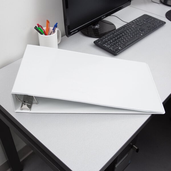 A white Cardinal tabloid size binder with a computer monitor and cup of pens on a white desk.