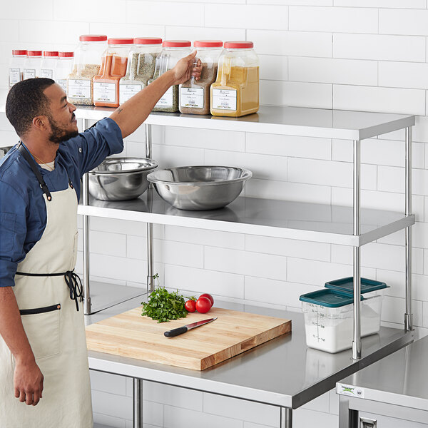 A man in an apron standing in a professional kitchen in front of a Regency stainless steel double deck overshelf.