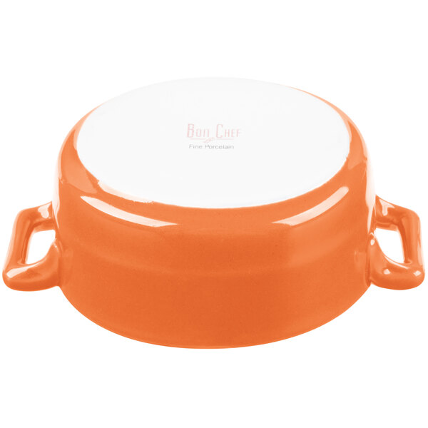 An orange porcelain oval cocotte with white handles.
