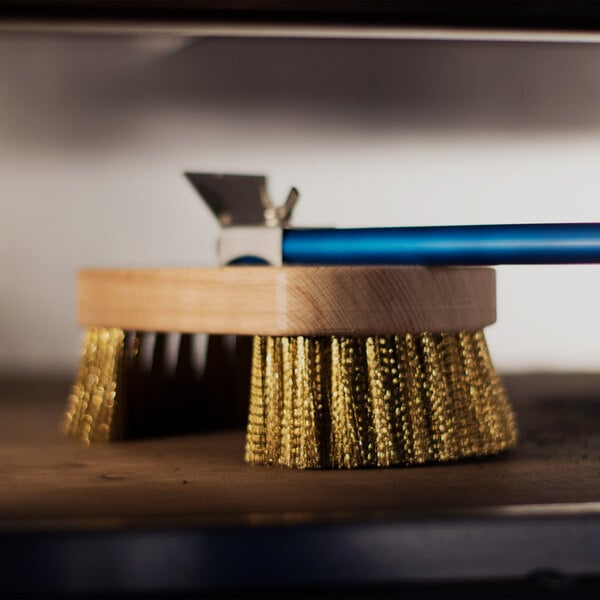 A round brush with brass bristles, a wooden block, and a blue handle, featuring a metal scraper attachment.