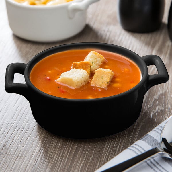 A matte black porcelain oval cocotte filled with soup and croutons.