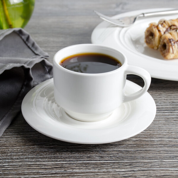 A cup of coffee on a Bon Chef white porcelain demi saucer on a table with food.