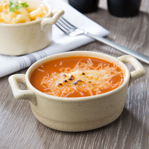 A close up of a bowl of soup with cheese and pasta in a Bon Chef wheat porcelain cocotte.