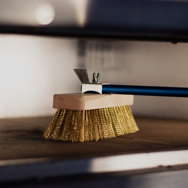 A brass-bristled brush with a wooden block and a blue handle is being used to clean the surface inside an oven.