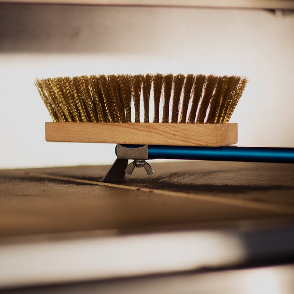 A close-up of a brass-bristled brush with a wooden block and a blue handle, resting on a flat surface.