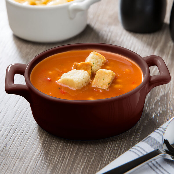 An oval burnt umber porcelain dish filled with soup and croutons.