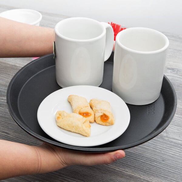 A person holding a black Cambro non-skid serving tray with a plate of cookies and two white mugs.