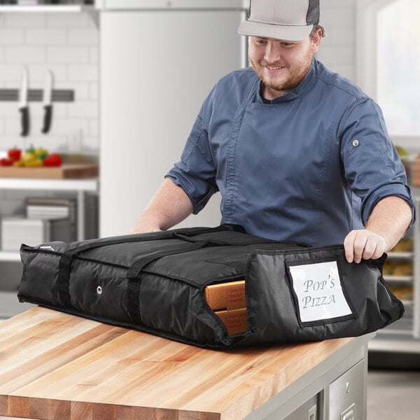 A man in a chef's hat holding a black CaterGator Dash pizza bag with brown pizza boxes inside.