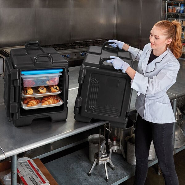 A woman in a white coat and gloves using a black Cambro front loading food pan carrier to hold a tray of food.