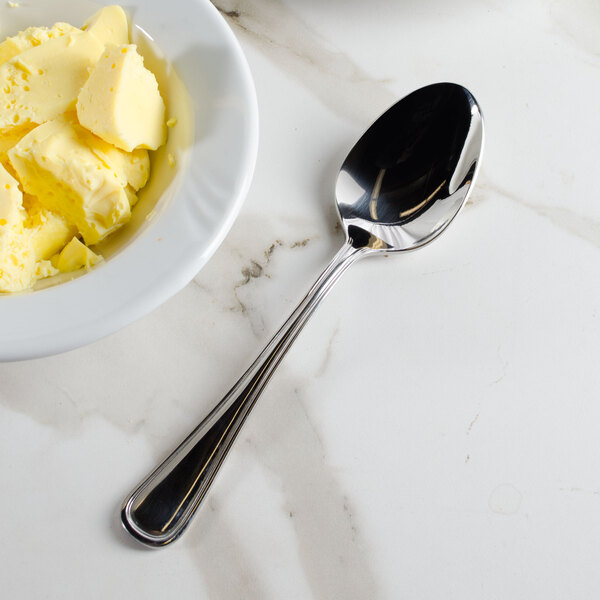 A Walco Pacific Rim stainless steel serving spoon in a bowl of butter on a marble surface.