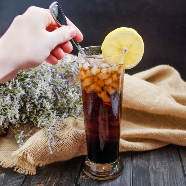 A hand holding a Walco Luxor stainless steel iced tea spoon over a glass of ice tea with a lemon slice.
