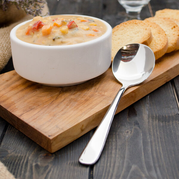 A Walco 18/10 stainless steel bouillon spoon on a wood board next to a bowl of soup.