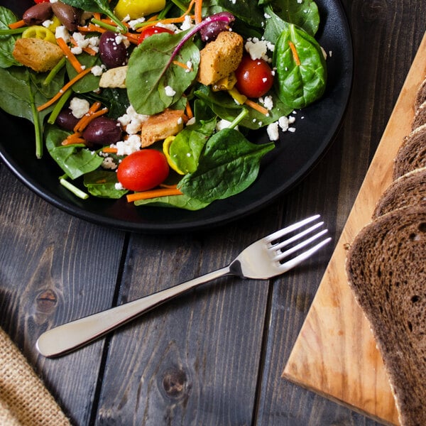 A plate of salad with a Walco Star stainless steel salad fork and bread on a wood table.