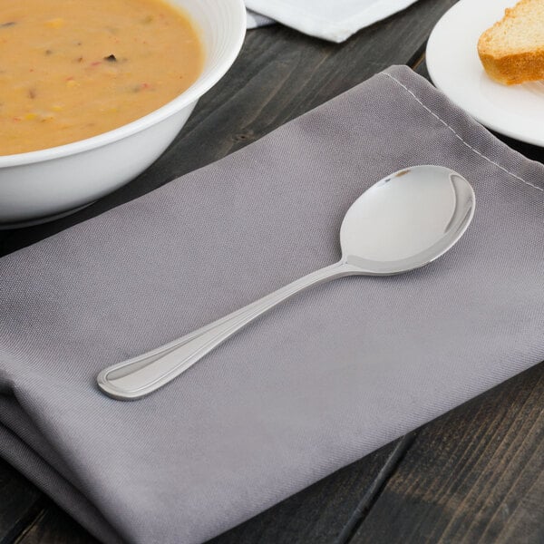 A Walco Pacific Rim stainless steel bouillon spoon on a napkin next to a bowl of soup.