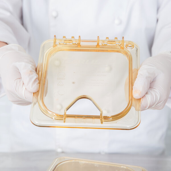 A person in white gloves holding a Carlisle amber plastic container with a hinged lid.