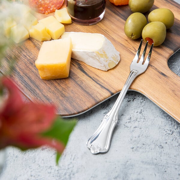 A Walco Barony stainless steel cocktail fork on a cutting board with cheese and olives.