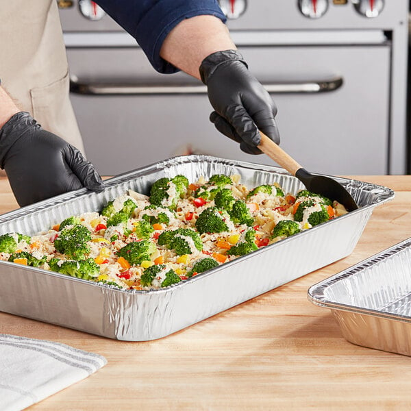 A gloved hand cutting broccoli and cheese in a Western Plastics foil steam table pan.