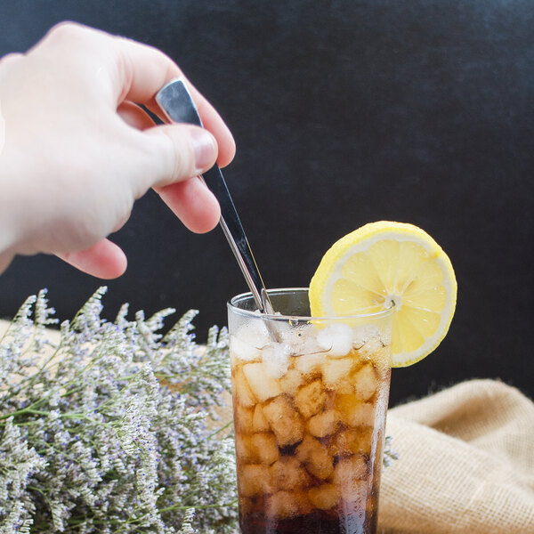 A hand holding a Walco Freya stainless steel iced tea spoon over a glass of iced tea with a lemon slice.