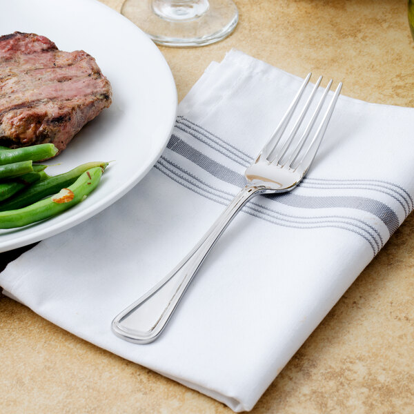 A Walco stainless steel table fork on a napkin next to a plate of meat and green beans.