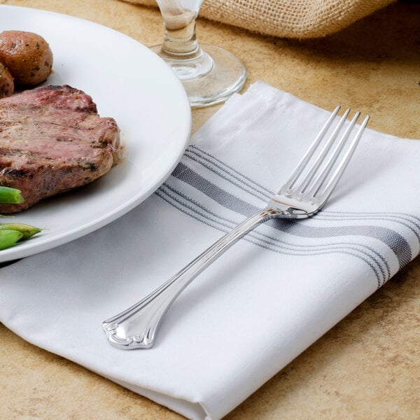 A Walco stainless steel dinner fork on a napkin next to a plate of meat and vegetables.