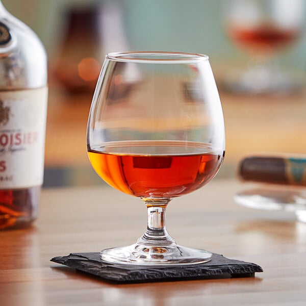 A customizable Acopa brandy snifter filled with brown liquid on a coaster on a table.