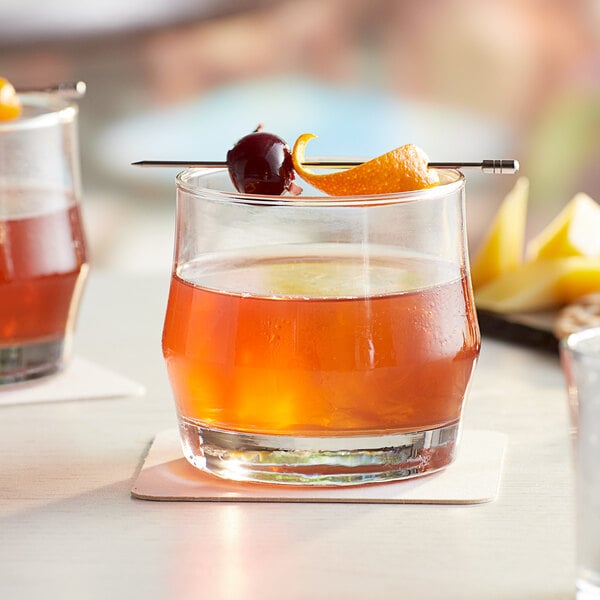 Two Acopa old fashioned glasses with brown cocktails and fruit garnish on a table.