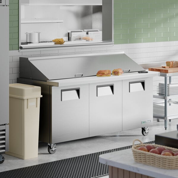 A stainless steel, three-door mega top refrigerated sandwich prep table with a raised ingredient rail and cutting board.