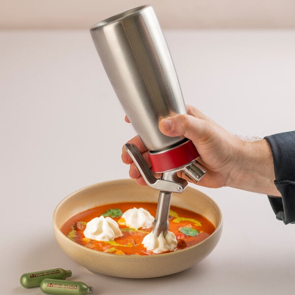 A hand is using a stainless steel whipped cream dispenser to add dollops of cream to a bowl of soup, with two small gas cartridges visible nearby.