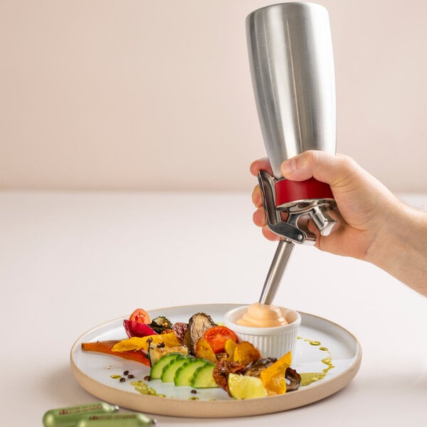 A hand is using a stainless steel whipped cream dispenser to dispense a creamy topping into a small dish on a plate of assorted vegetables.
