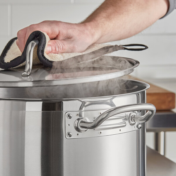 A large stainless steel sauce pot with a lid being lifted by a hand using an oven mitt.