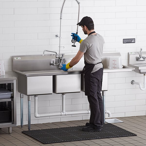 A man in a black shirt and blue pants using a Regency stainless steel three compartment sink in a professional kitchen.