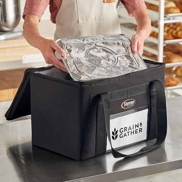 A person putting a tray of food into a Sterno black insulated delivery bag.