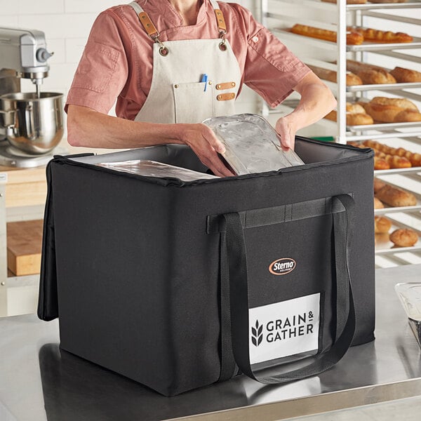 A woman wearing an apron putting food in a Sterno extra-large black nylon food carrier bag.