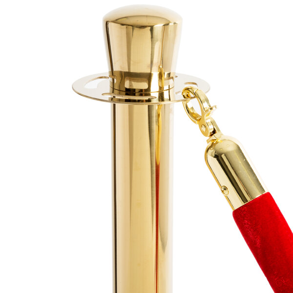A close-up of a gold Lancaster Table & Seating crowd control stanchion pole with red velvet rope.