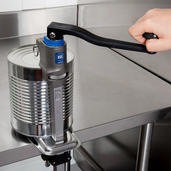 A heavy-duty can opener with a long handle mounted on a metal countertop, being used to open a large can.
