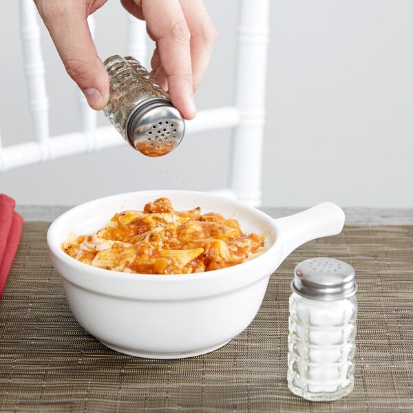 A hand using a Tablecraft Nostalgia glass salt shaker to add salt to a bowl of food.
