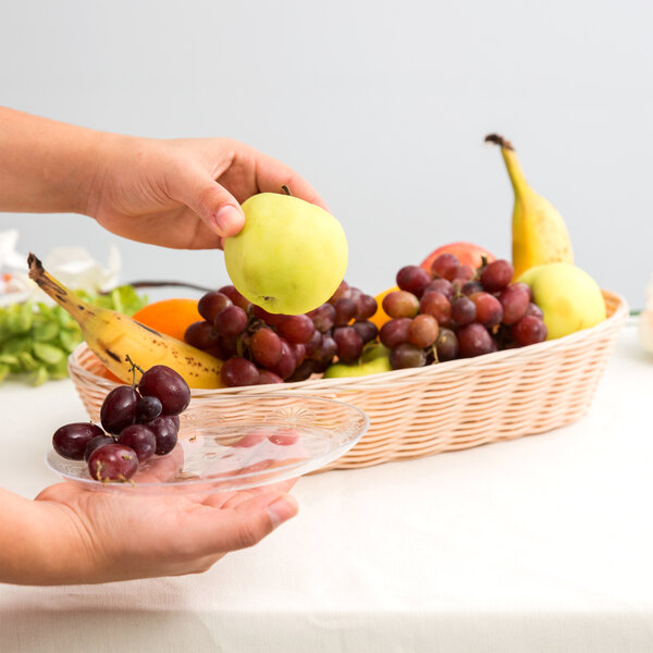 A hand holding a Tablecraft woven rattan-like bread basket over a plate of fruit.