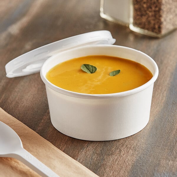 A white paper food cup of soup with a white plastic lid and a spoon on a table.