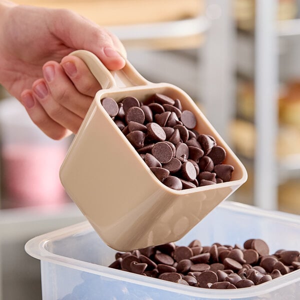 A hand using a Carlisle beige portion scoop to pour chocolate chips into a container.