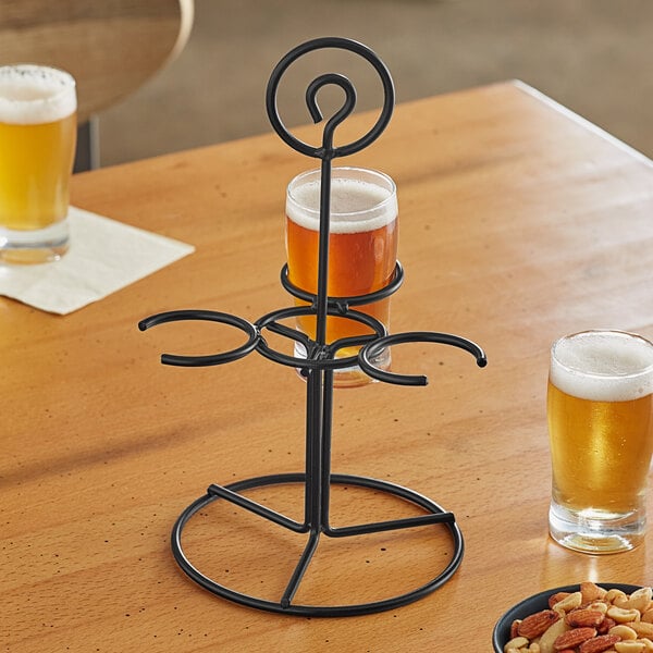 An Acopa black metal flight carrier with three glasses of beer and a plate of nuts on a table.