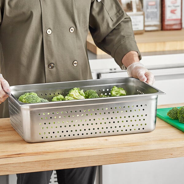 A man in a chef's coat holding a Choice stainless steel steam table pan filled with broccoli.