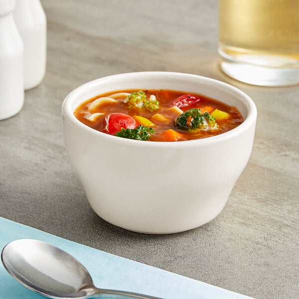 An Acopa ivory stoneware bouillon cup filled with soup with vegetables and a spoon.