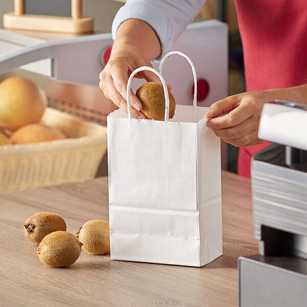 A person holding a kiwi and putting it in a white paper shopping bag.