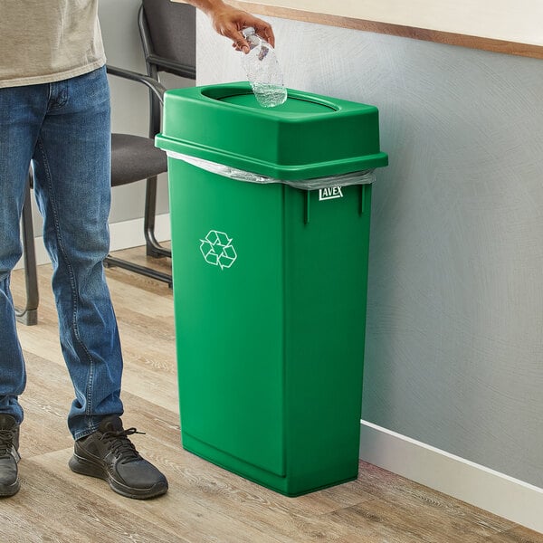 A person throwing a plastic bottle into a green Lavex slim rectangular recycling bin.