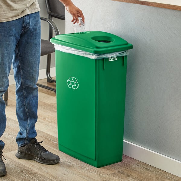 A person putting a plastic bag into a Lavex green rectangular recycle bin with a green lid.