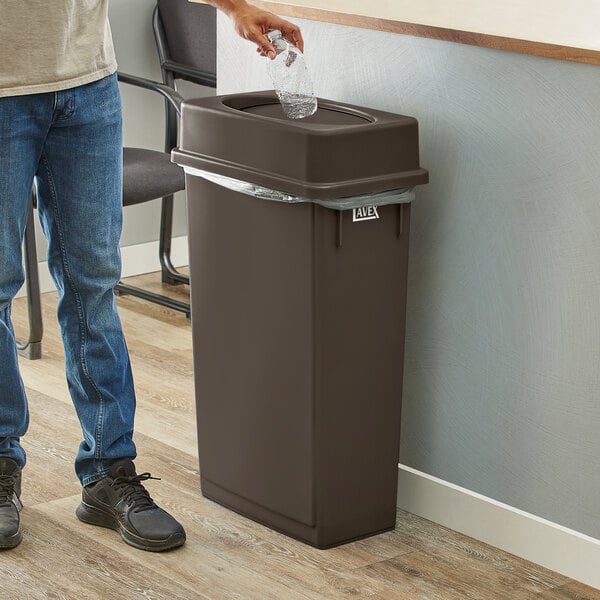 A man in a corporate office cafeteria throws a plastic bottle into a brown Lavex Slim Rectangular Trash Can with a brown Drop Shot Lid.